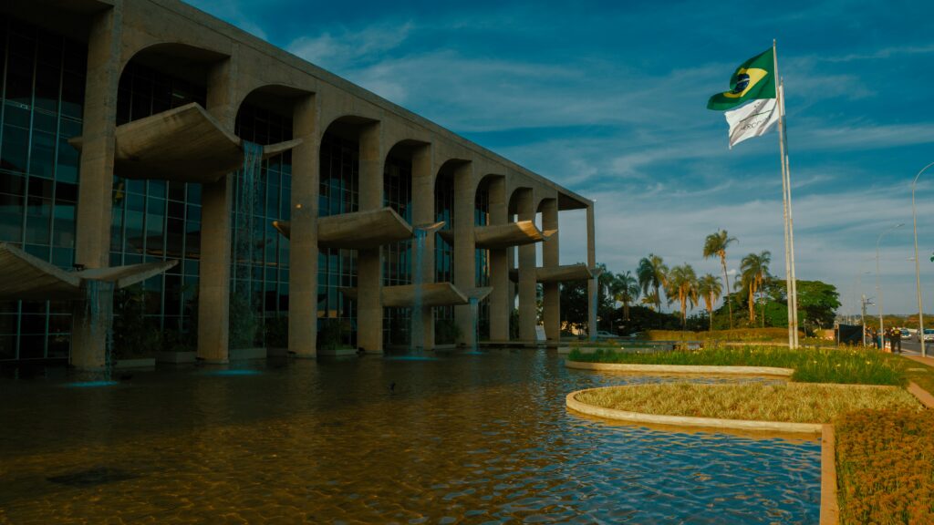 A modern building with large arches stands beside a reflective pool, with the Brazilian flag and another white flag waving on flagpoles nearby, palm trees and a blue sky in the background.