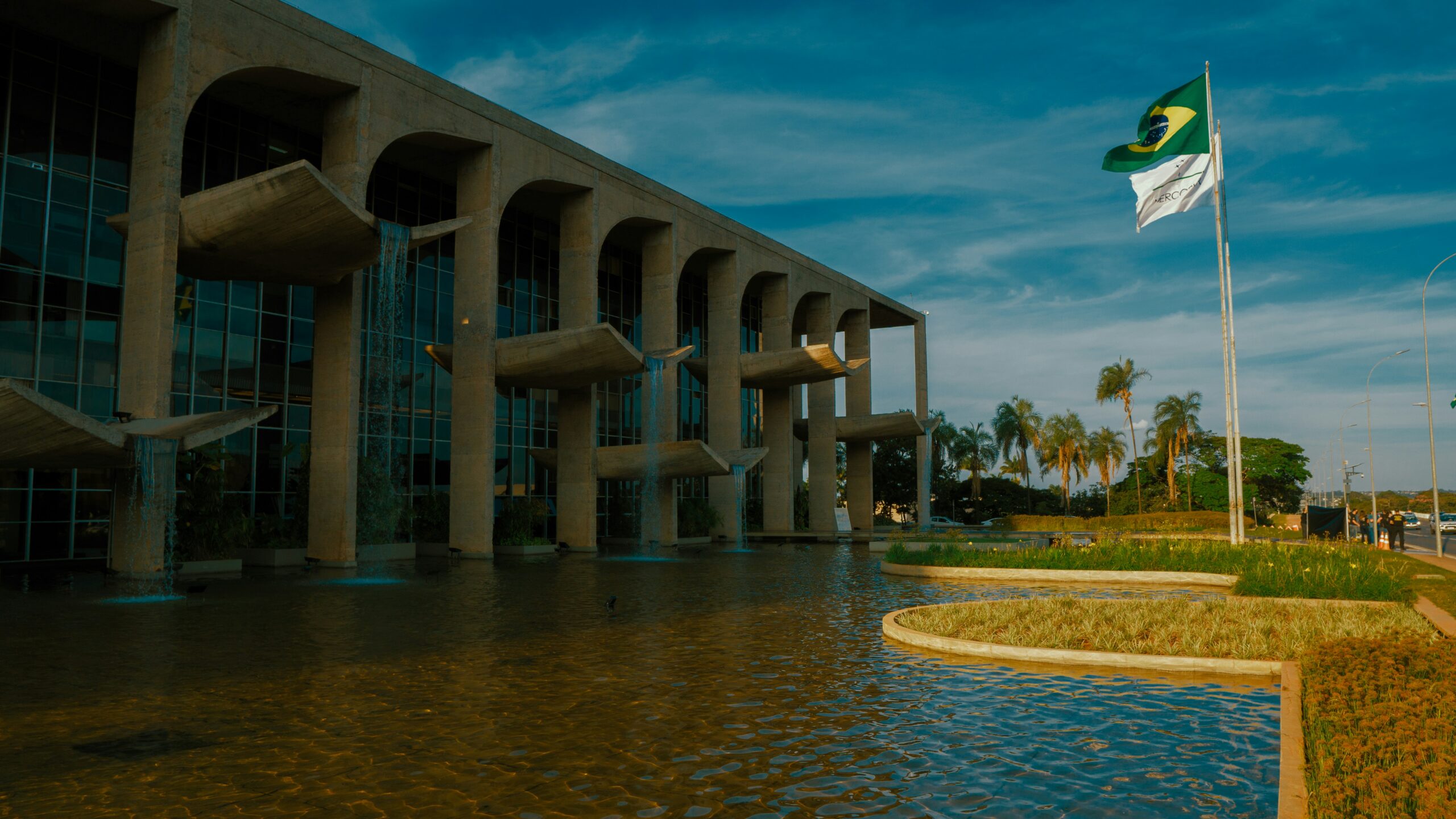 A modern building with large arches stands beside a reflective pool, with the Brazilian flag and another white flag waving on flagpoles nearby, palm trees and a blue sky in the background.
