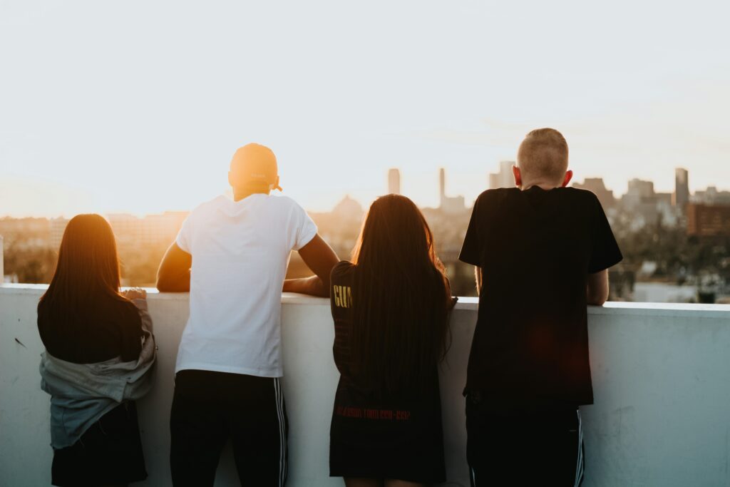 Four people stand side by side, leaning on a white wall, and look out over a city skyline at sunset, with the sun glowing brightly in the sky ahead of them.