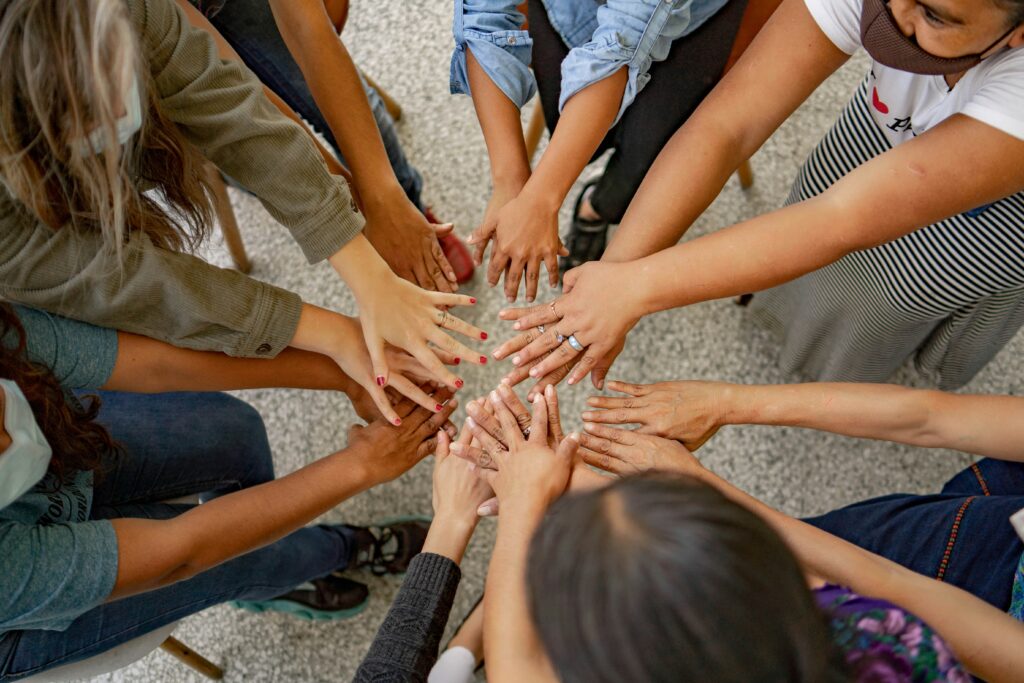 A group of people in a circle, with their hands stacked in the center. Their hands are varied in skin tone, with some wearing rings and nail polish.