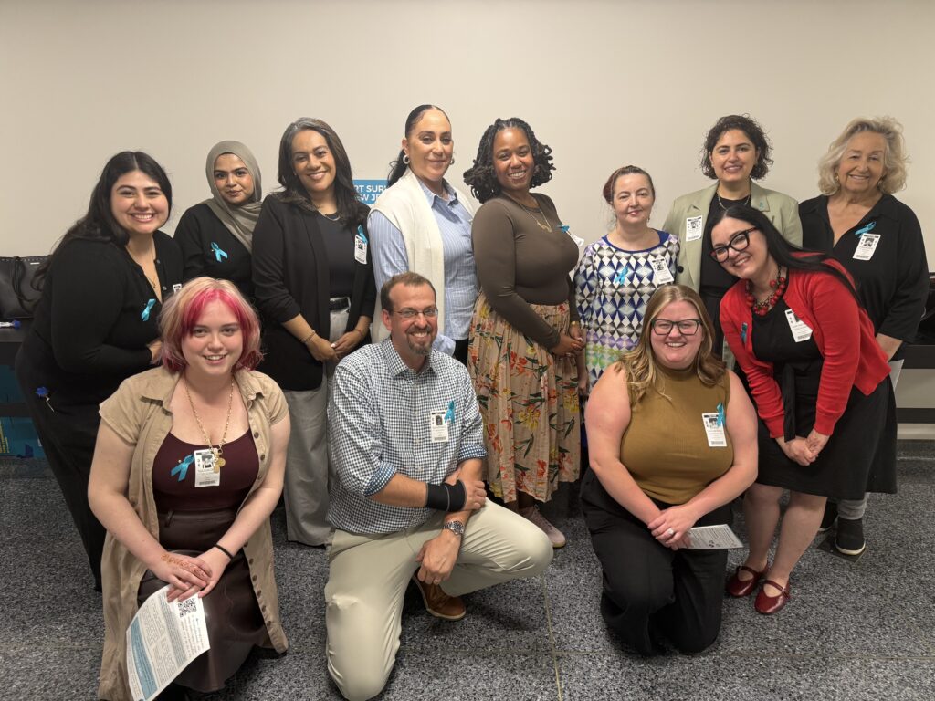 A group of twelve professionally dressed adults, diverse in gender and ethnicity, pose and smile together indoors against a plain wall. Several wear teal awareness ribbons and hospital or staff badges.
