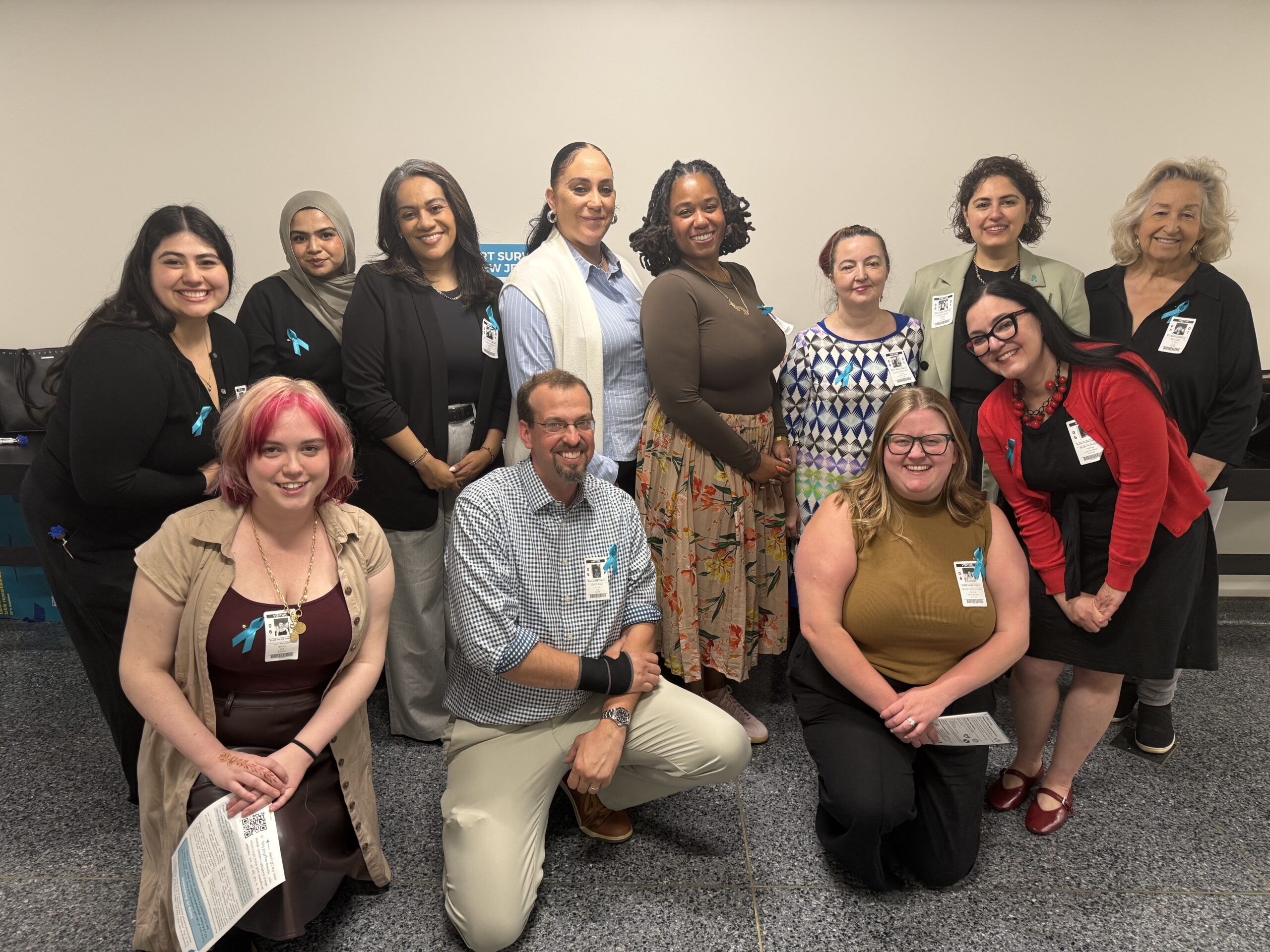 A group of twelve professionally dressed adults, diverse in gender and ethnicity, pose and smile together indoors against a plain wall. Several wear teal awareness ribbons and hospital or staff badges.
