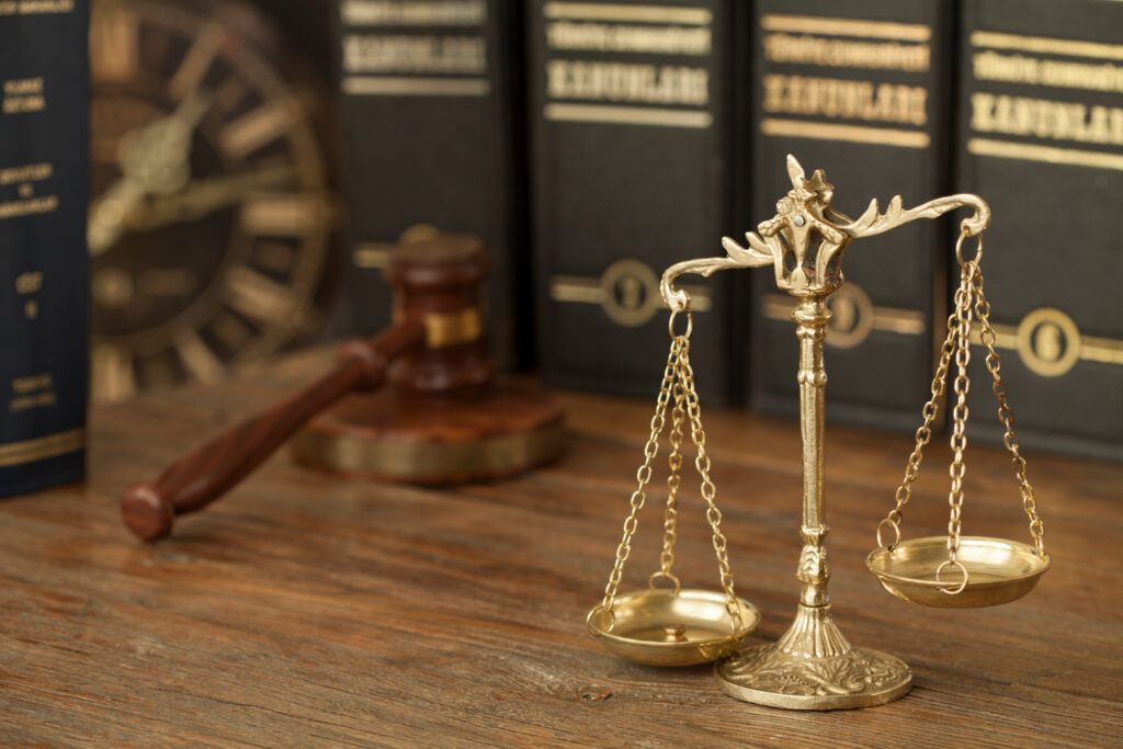 A brass scale of justice standing on a wooden desk in front of a judges gavel and law books.